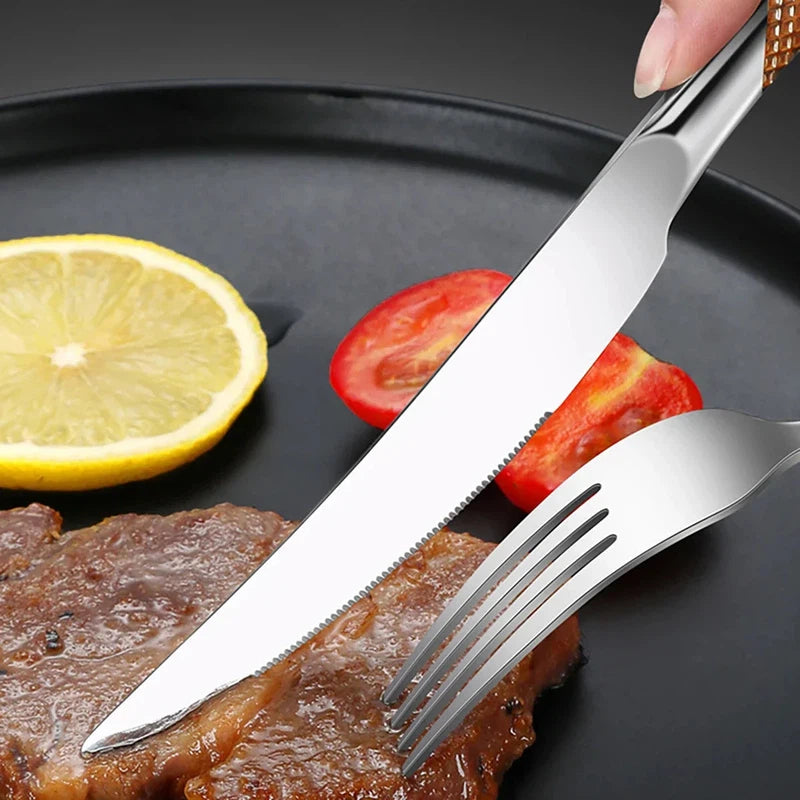 Stainless steel knife and fork from 24pcs tableware set cutting steak on black plate with lemon and tomato slices.