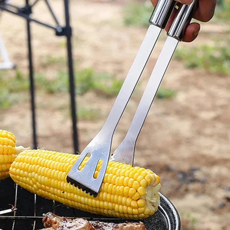 Stainless steel barbecue tools in use, grilling corn and meat outdoors.