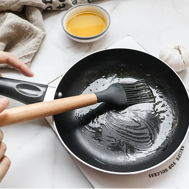 Non-stick silicone spatula with wooden handle applying oil to a frying pan, part of a 12-piece kitchen utensil set.